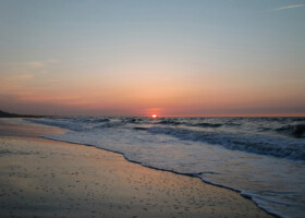 Blick auf einen Strand und das Meer auf dem die Sonne gerade untergeht und den Horizont rot färbt